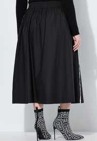Femme portant une jupe midi noire à taille élastique et des bottines à talons hauts à motifs géométriques noir et blanc, se tenant contre un mur blanc.