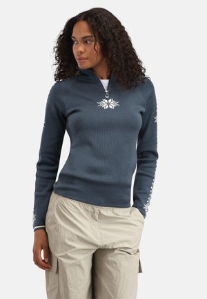 Kvinne med krøllete hår iført en mørk blå genser med kvartglidelås og hvite snøkrystallmønstre og beige cargobukser, som ser til siden.