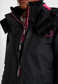 Schwarze wasserdichte Jacke mit pinken Reißverschlüssen und Akzenten, strukturiertem Material, hohem Kragen und einem Klettverschluss-Patch auf der Schulter.
