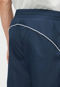 Κοντινό πλάνο ενός ατόμου που φοράει σορτς ναυτικού μπλε με λευκή διακοσμητική ραφή και ελαστική μέση, σε συνδυασμό με ένα ανοιχτό γκρι πουκάμισο.