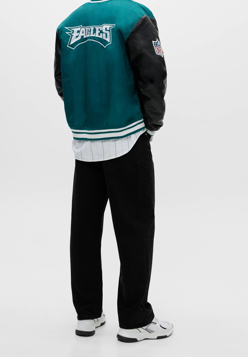 Personne portant une veste Eagles NFL verte et noire, un pantalon noir, des baskets blanches et une chemise blanche à rayures verticales, debout dos à nous.