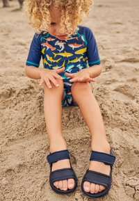 Sandali blu navy con due cinturini regolabili in velcro, posizionati su un terreno sabbioso. Un bambino in un costume da bagno colorato a fantasia squalo è seduto nelle vicinanze.