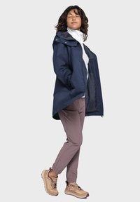 Marineblauer wasserdichter Mantel mit Kapuze, weißer Innenlage und hellrosa Hose. Beigefarbene Sneaker mit strukturierten Sohlen und dunklen Akzenten.