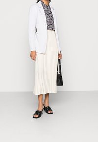 Frau trägt einen weißen Blazer, eine schwarz-weiße gemusterte Bluse, einen plissierten cremefarbenen Midirock, schwarze Absatzsandalen und hält eine schwarze Handtasche.