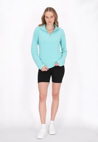 Lyseblå langærmet pullover med en kvart lynlås, parret med sorte shorts og hvide sneakers. Stoffet ser glat og struktureret ud.
