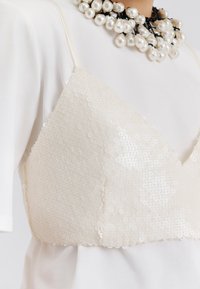 Ziloņkaula krāsas sequinēta bralete ar trīsstūra formu un regulējamām lencēm, pārslāņota pār vienkāršu baltu blūzi. Dekorēta ar pērļu kaklarotu.