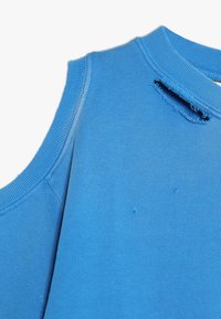 Felpa senza maniche blu con tessuto texture, scollatura a costine e dettagli usurati vicino alla spalla e all'orlo.