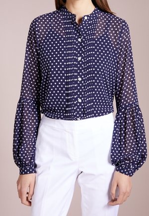 Granatowa przezroczysta bluzka w białe kropki, zapięcie na guziki z przodu, plisowany detal oraz bufiaste długie rękawy, zestawiona z białymi spodniami.
