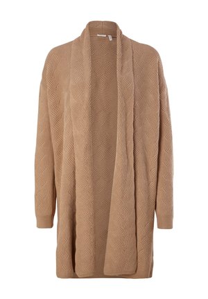 Mujer con suéter de cuello alto beige, cárdigan largo marrón, pantalones ajustados crema y botines crema con detalle de cremallera.