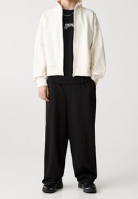 Weiße Fleecejacke mit Reißverschluss über einem schwarzen T-Shirt mit grafischem Druck, kombiniert mit lockeren schwarzen Hosen und schwarzen Sneakern mit strukturiertem Design.