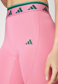 Różowe legginsy sportowe o gładkiej teksturze, z zielonym i różowym paskiem w talii oraz zielonym logo z boku.