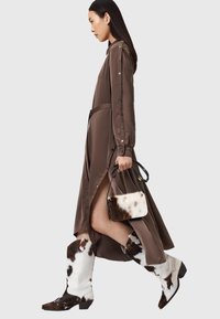 Abito marrone a maniche lunghe con bottoni decorativi e cintura in vita, abbinato a stivali bianchi e marroni con stampa a motivo mucca e una borsa coordinata.