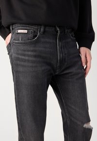 Jeans en denim noir avec une coupe décontractée. Les caractéristiques incluent une poche avant, une texture délavée et une légère déchirure au genou droit. Étiquette Calvin Klein visible.