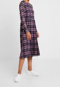 Vestido de manga larga a cuadros en negro y morado con cintura fruncida y bolsillos, combinado con zapatillas de plataforma blancas. Textura de tela suave.