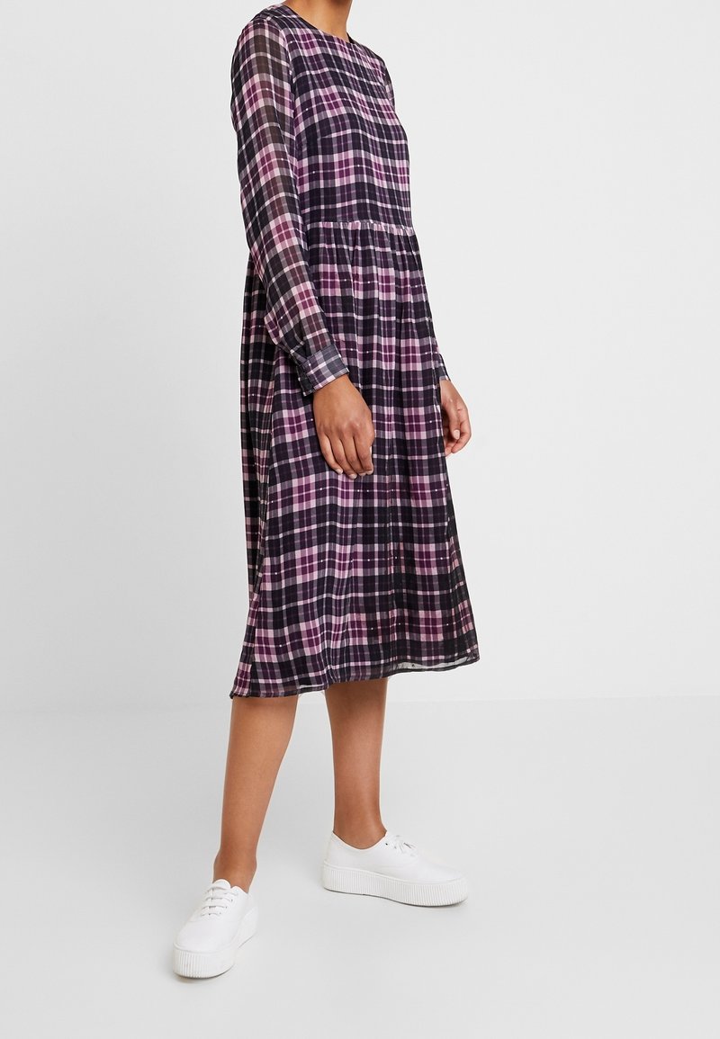 Vestido de manga larga a cuadros en negro y morado con cintura fruncida y bolsillos, combinado con zapatillas de plataforma blancas. Textura de tela suave.