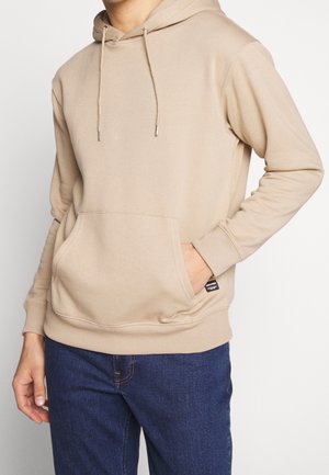 Mann trägt beigen Kapuzenpullover mit Fronttasche und blaue Jeans, Hand in die Tasche gesteckt vor einem schlichten weißen Hintergrund.