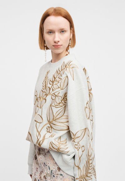 Νεαρή γυναίκα με κοντά κόκκινα μαλλιά, που φοράει μεγάλα σκουλαρίκια με πέρλες, ένα ανοιχτό φούτερ με χρυσή κεντητή ανθοδέσμη και μια φλοράλ φούστα.