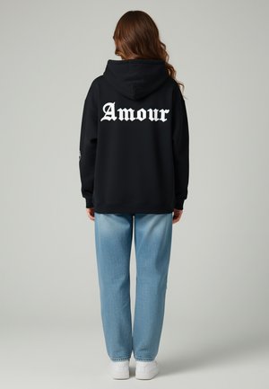 Felpa nera con cappuccio e testo bianco "Amour" sulla schiena, abbinata a jeans azzurri e sneakers bianche, su uno sfondo neutro.