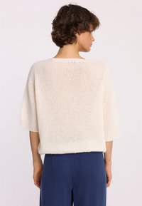 Osoba z krótkimi kręconymi włosami, ubrana w luźny biały sweter dzianinowy i ciemnoniebieskie spodnie, zwrócona tyłem w stronę jednolitego jasnego tła.