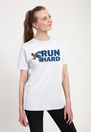 Junge Frau mit langem Pferdeschwanz, die ein weißes T-Shirt mit der Grafik eines Fußballspielers und dem Text "RUN HARD" trägt, steht vor einem schlichten Hintergrund.