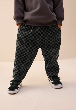 Pantaloni de trening în carouri negre și gri, cu o talie elastică și buzunare laterale, purtați cu adidași cu velcro negri și albi.