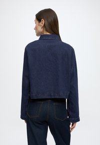 Kort mørk denimjakke med lange ærmer, en krave og synlige sømme; båret med mørke jeans med udvendige baglommer.