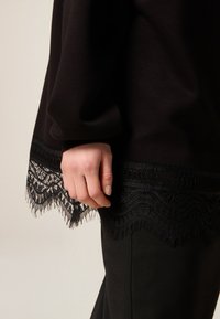 Top negro con un borde de encaje en la parte inferior. El encaje tiene un borde festoneado y patrones intrincados. Mano descansando cerca del dobladillo.