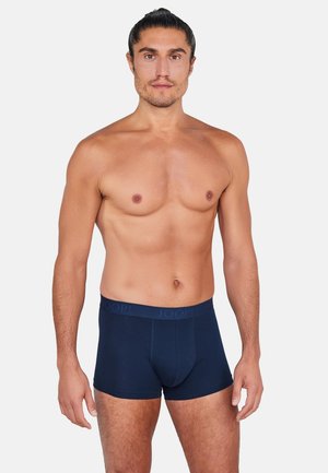 Mężczyzna bez koszulki z włosami związanymi do tyłu, ubrany w ciemnoniebieskie bokserki, stojący na jednolitym białym tle.