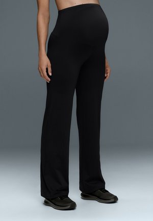 Personne enceinte portant un pantalon de maternité noir et des baskets noires, debout devant un fond gris uni.