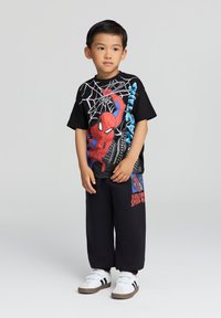 T-shirt nera con grafica di Spider-Man in rosso e blu, abbinata a pantaloni neri con un'accenno stampato, completata da sneakers bianche.