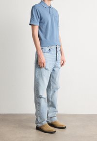 Niebieska bawełniana koszulka polo z krótkim rękawem, zestawiona z jasnymi, postarzanymi jeansami i brązowymi butami wsuwanymi.