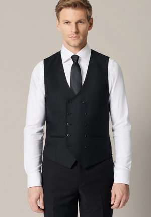 Mężczyzna w białej koszuli, czarnej kamizelce dwurzędowej, czarnym krawacie i czarnych spodniach, stojący na tle jednolitego beżowego tła.