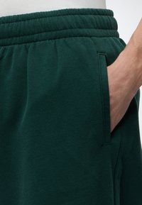Main dans la poche d’un pantalon de jogging vert foncé à taille élastique porté avec un haut blanc, montrant la poche de couture latérale et la texture du tissu.
