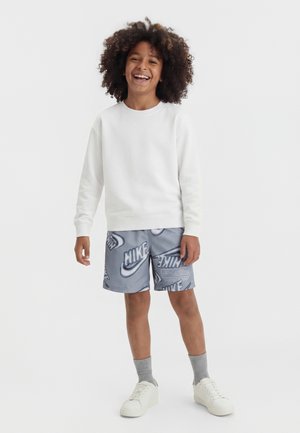 Barn med krøllet hår iført en hvid sweatshirt, grå Nike-logo shorts, grå sokker og hvide sneakers, smilende og stående på en hvid baggrund.