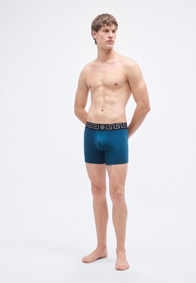 Τιρκουάζ ανδρικά εσώρουχα μποξεράκια με μαύρη ζώνη μέσης με σχέδια. Το ύφασμα φαίνεται λείο και ελαστικό. Το μοντέλο στέκεται ξυπόλυτο, με τα χέρια χαλαρά στο πλάι.