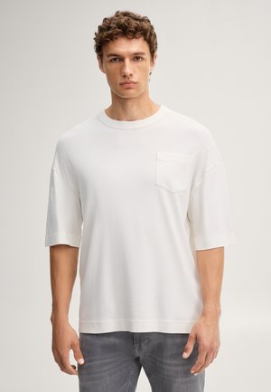 Junger Mann mit lockigem Haar trägt ein schlichtes weißes übergroßes T-Shirt mit Brusttasche und graue Jeans, steht vor hellem Hintergrund.