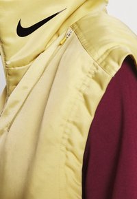 Gul vadderad väst med huva, med en svart Nike-logotyp, sidodragkedja och kontrasterande maroon inredning. Slät, blank textur.