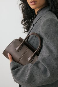 Brązowa skórzana torebka na ramię z dwoma okrągłymi uchwytami, prostokątnym kształtem i przeszyciami. Nosi ją w komplecie ze szorstką szaro-skalą marynarką.
