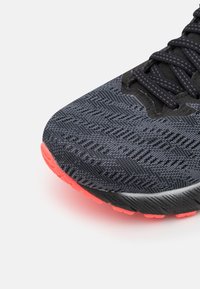 Primer plano de una zapatilla de running negra de punto con cordones oscuros y suela de goma roja brillante sobre un fondo blanco.
