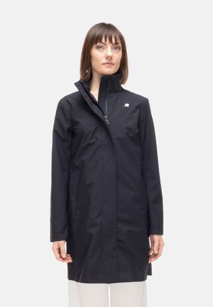 Donna con impermeabile nero al ginocchio con collo alto, logo bianco sul petto e pantaloni bianchi su sfondo chiaro uniforme.