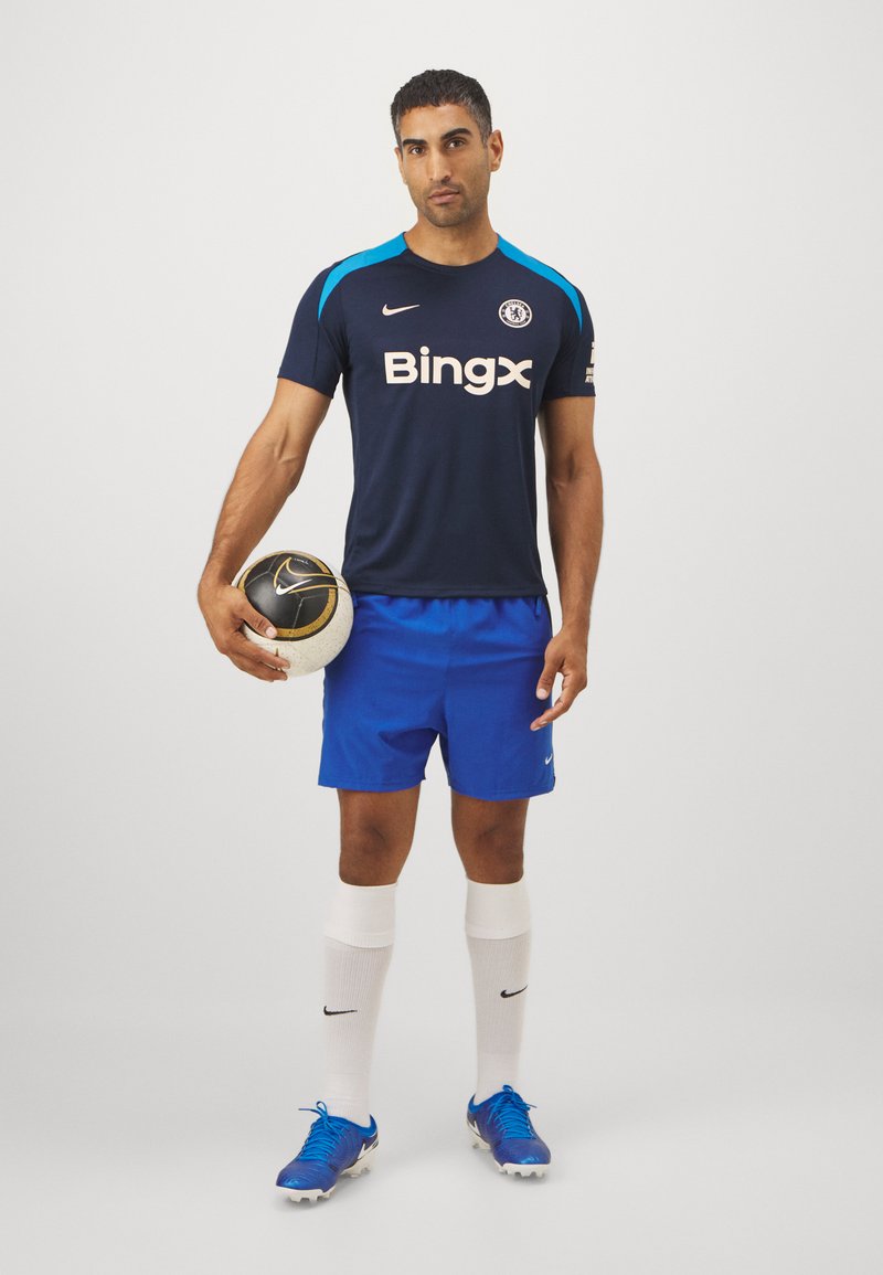 Niebieska koszulka piłkarska z czarnymi i turkusowymi akcentami, białym logo "Bingx", w zestawie z niebieskimi szortami, białymi skarpetkami i niebieskimi spodniami do piłki nożnej, trzymająca piłkę.