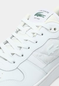 Λευκό δερμάτινο αθλητικό παπούτσι με υφή, διαθέτει λευκά κορδόνια, λεπτό ανάγλυφο σχέδιο και πράσινο λογότυπο στη γλώσσα.