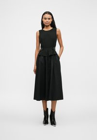 Μαύρο αμάνικο φόρεμα με εφαρμοστό κορσέ, μέση πιέτα και φούστα σε γραμμή Α. Διαθέτει μεταλλικές λεπτομέρειες και λεία υφή.