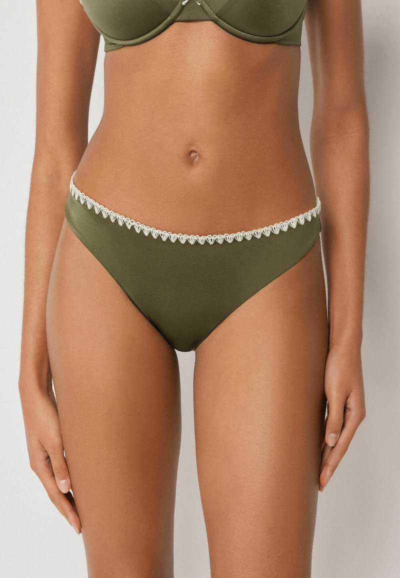 Frau trägt olivgrüne Bikinihose mit weißem, wellenförmigem Saum, zeigt Bauch und obere Oberschenkel vor einfachem Hintergrund.