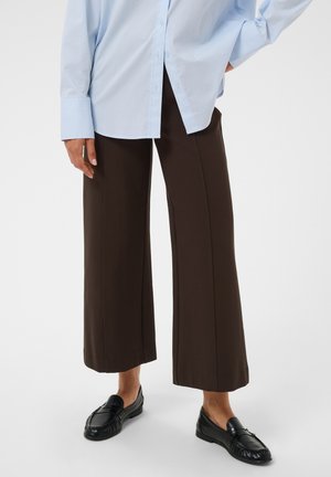 Personne portant un pantalon marron large et court, une chemise bleu clair à manches longues et des mocassins en cuir noir, debout avec une main sur la hanche.