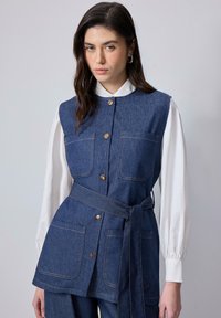 Gilet en denim au design structuré, doté de deux poches avant et d'une taille nouée, associé à une chemise blanche à manches longues. Comprend des boutons dorés.