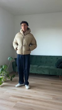 Μπεζ φουσκωτό μπουφάν με ψηλό γιακά και κλείσιμο με φερμουάρ, συνδυασμένο με σκούρα παντελόνια σε φαρδιά γραμμή και λευκά αθλητικά παπούτσια. Εσωτερικός χώρος με έναν πράσινο καναπέ.