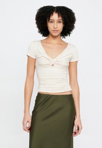 Világos bézs színű, testhezálló felső rövid ujjakkal, V-nyakkivágással, elöl csavart részlettel és oldalán ráncolással, sötét olívazöld szoknyával párosítva.