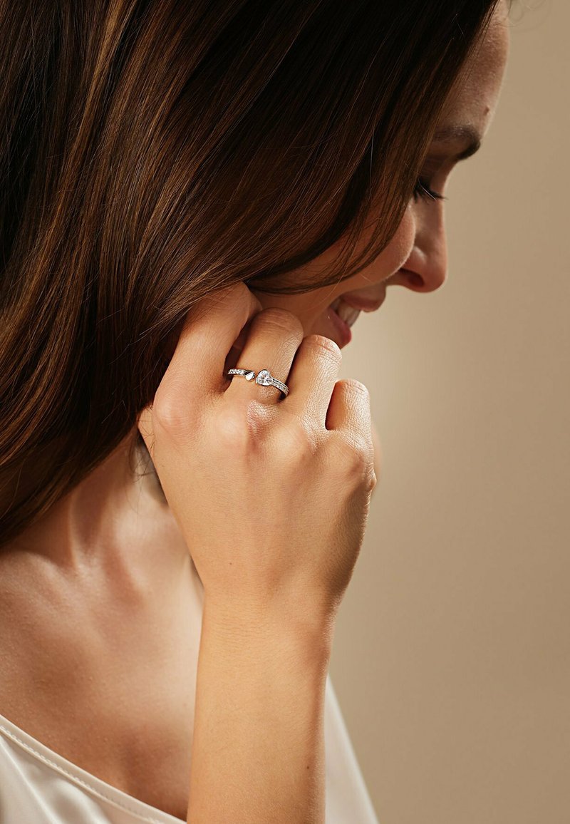 Sølvring med et hjerteformet design, prydet med små klare sten langs bæren, båret på en hånd med glat hud mod en neutral baggrund.