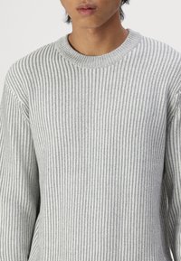 Pull en tricot gris à côtes avec un col rond. Présente un tissu texturé et des manches longues. Silhouette ajustée sans ornements.